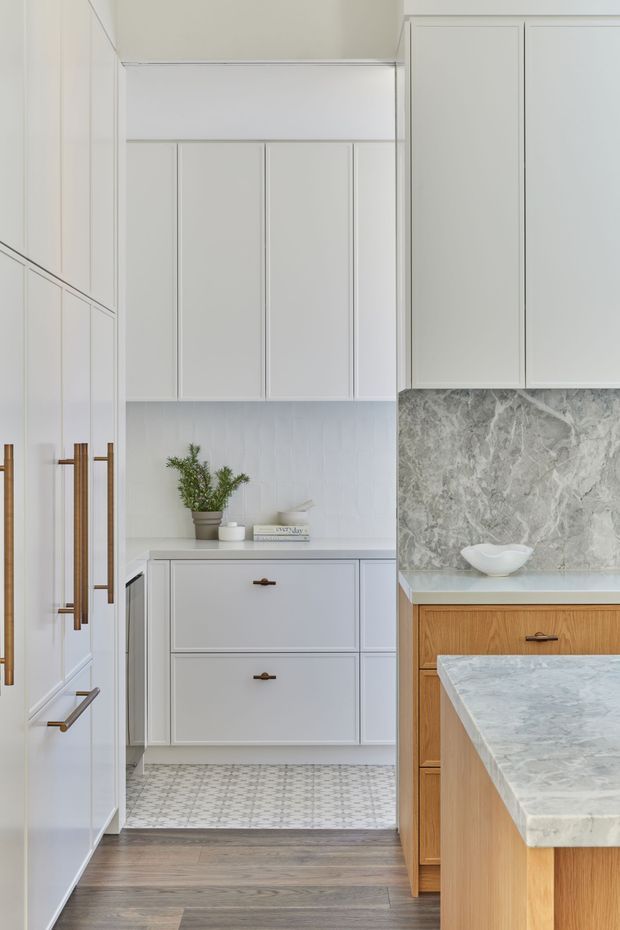 A bright, refined kitchen featuring extensive white shaker-style cabinetry, slim brass hardware, a grey veined stone splashback and benchtop, warm timber island cabinetry, and a patterned tiled floor transitioning to dark timber flooring. A small pantry or secondary preparation area is visible beyond the main kitchen.