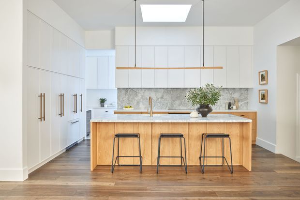 A bright, spacious contemporary kitchen with a large timber island and three black bar stools. White full-height cabinetry lines the walls, with integrated appliances, a pale stone benchtop, and a grey-veined stone splashback. A linear timber pendant hangs above the island, while a skylight provides additional natural light. Warm timber flooring contrasts with the crisp white joinery.
