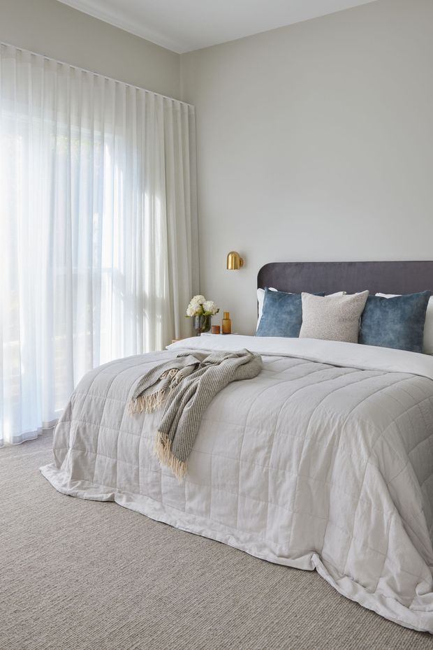 A softly styled contemporary bedroom with a large upholstered charcoal-grey bedhead, white quilted bedding, muted blue and cream cushions, and a textured grey throw. Full-height sheer curtains allow abundant natural light into the room, while a small brass wall sconce and bedside styling add warmth. The room has neutral painted walls and wall-to-wall grey carpet.