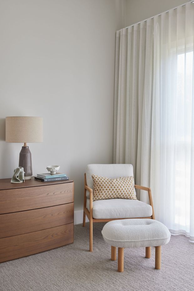A softly lit, minimalist sitting corner with a mid-century-inspired timber three-drawer dresser, textured table lamp, upholstered wooden armchair, patterned cushion, and matching ottoman. Floor-to-ceiling sheer curtains and heavier drapes frame a large window, while neutral walls, carpet, and upholstery create a calm, warm interior.