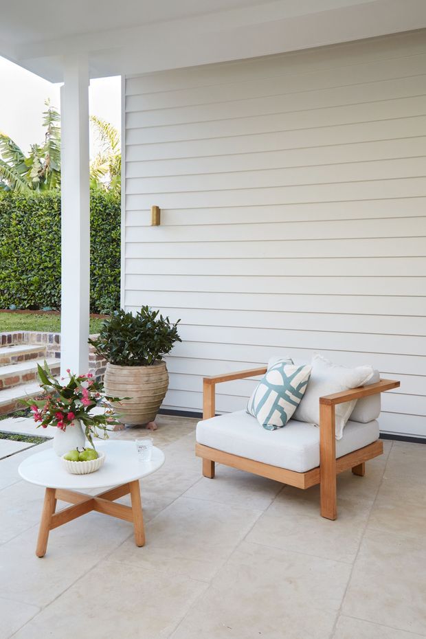 A bright covered outdoor sitting area with a white horizontal weatherboard wall, pale tiled floor, and a timber-framed lounge chair with white cushions and a patterned blue accent pillow. A small round timber-and-white coffee table holds a vase and bowl, while a large potted plant and landscaped garden are visible beside brick steps. The space has a relaxed, refined coastal feel.