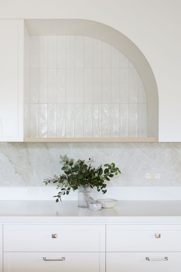 A bright, refined kitchen detail featuring white built-in cabinetry with brushed metal handles, a pale stone benchtop and matching stone splashback, and a glossy white vertically tiled arched wall niche. A timber ledge runs beneath the arch, while a vase of greenery and small ceramic vessels sit on the counter. The composition is light, calm, and softly textured.