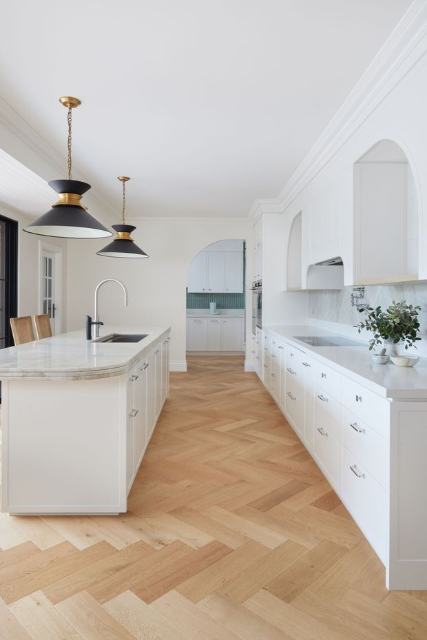 Bright, spacious white kitchen with extensive built-in cabinetry, a large island with integrated sink and curved stone benchtop, and a warm timber herringbone floor. The room features arched wall niches and openings, decorative ceiling cornices, black-framed glazing, and brass-and-black pendant lights. A secondary galley-style kitchen or pantry is visible through the rear archway.