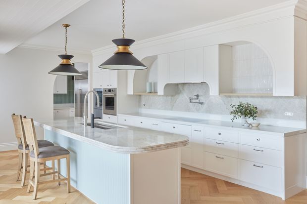 Bright, refined kitchen with extensive white built-in cabinetry, a large curved island with seating, pale stone surfaces and backsplash, arched open shelving, and two dark pendant lights. Herringbone timber flooring and ribbed island detailing add warmth and texture.