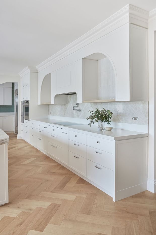 Bright, refined kitchen with extensive white built-in cabinetry, paneled fronts, a long stone benchtop and matching marble-look splashback. Large arched cabinetry and rangehood openings add a classical architectural detail, while brushed metal handles and a gooseneck tap provide subtle contrast. The floor is laid in a warm light timber herringbone pattern. A partial adjacent kitchen area is visible at left.