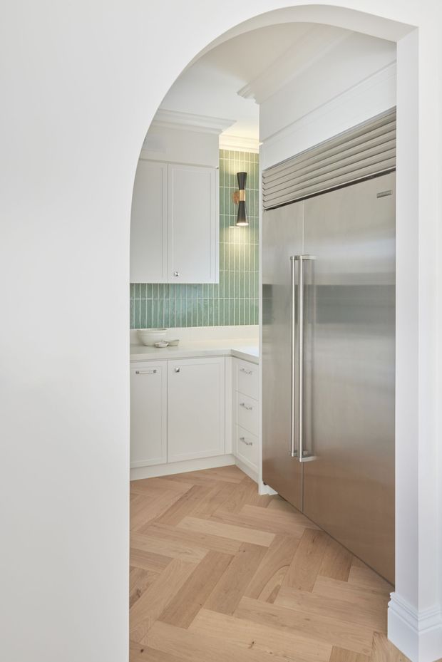 A compact, refined kitchen or butler's pantry viewed through a large white arched opening. The space features white shaker-style cabinetry, a pale green vertical tile splashback, a white stone benchtop, a large stainless-steel double-door refrigerator, and warm natural timber herringbone flooring. Decorative ceiling cornices, wall panelling, and a sculptural wall light add a traditional yet polished character.