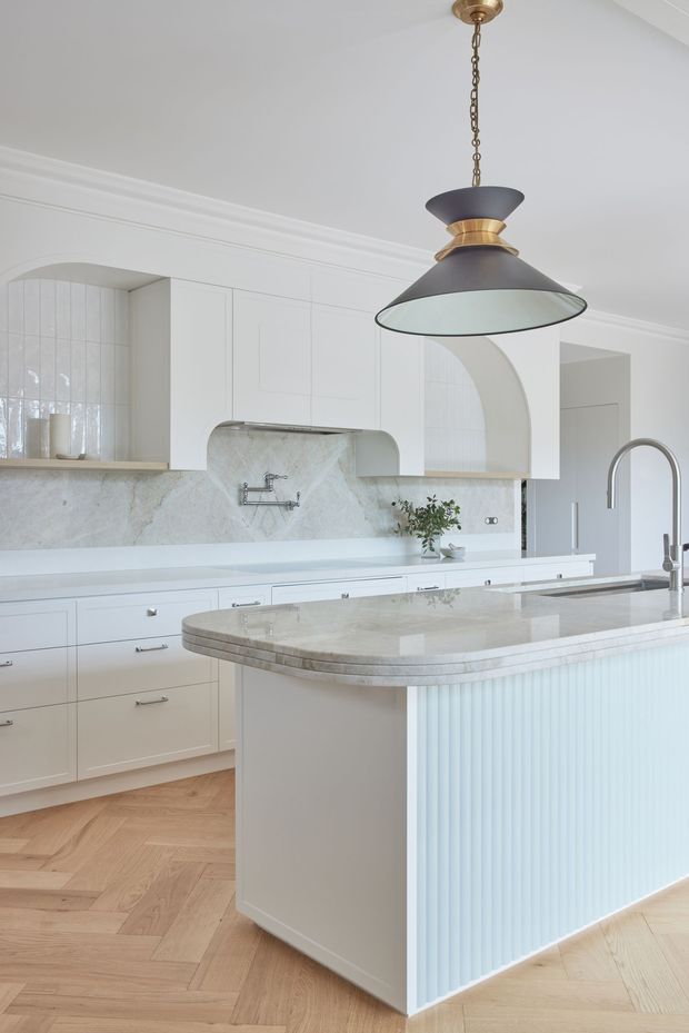 A bright, refined kitchen with extensive white cabinetry, a large marble or natural-stone island benchtop, pale fluted island panelling, and a timber herringbone floor. The kitchen features arched cabinetry and tiled niches, a high curved faucet, integrated appliances, and a prominent dark pendant light with brass detailing. Decorative ceiling mouldings and soft blue-white finishes create an elegant contemporary-classic feel.