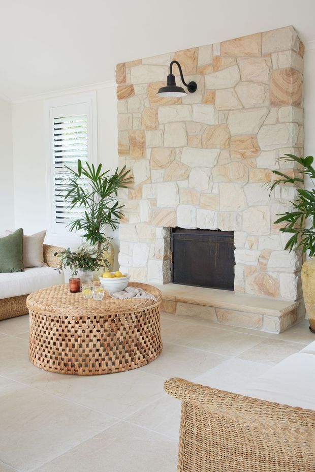 Bright, relaxed living room with a substantial irregular pale stone fireplace, black metal wall sconce and firebox, white plantation shutters, pale tiled flooring, woven rattan coffee table, wicker seating, and indoor palms. The room has a warm coastal and natural character.
