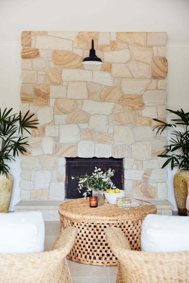 A bright, inviting living room centered on a large irregularly coursed natural stone fireplace with a dark firebox and a black pendant light above. White upholstered lounge chairs and a woven rattan coffee table occupy the foreground, with potted tropical plants flanking the fireplace and warm woven, natural-texture furnishings throughout.