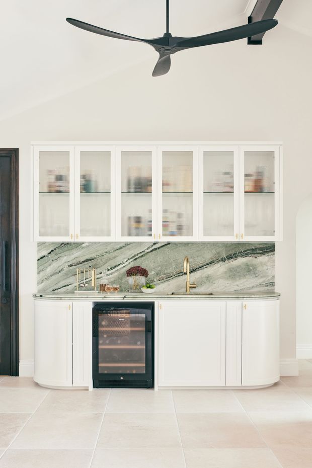 Bright, refined kitchen or entertaining joinery wall featuring white framed glass-front cabinetry, curved lower cabinets, a dramatic green-veined marble splashback and benchtop, brass tapware and hardware, and an integrated wine fridge. A sculptural dark ceiling fan hangs above, with pale large-format floor tiles and a dark timber-framed doorway at the side.