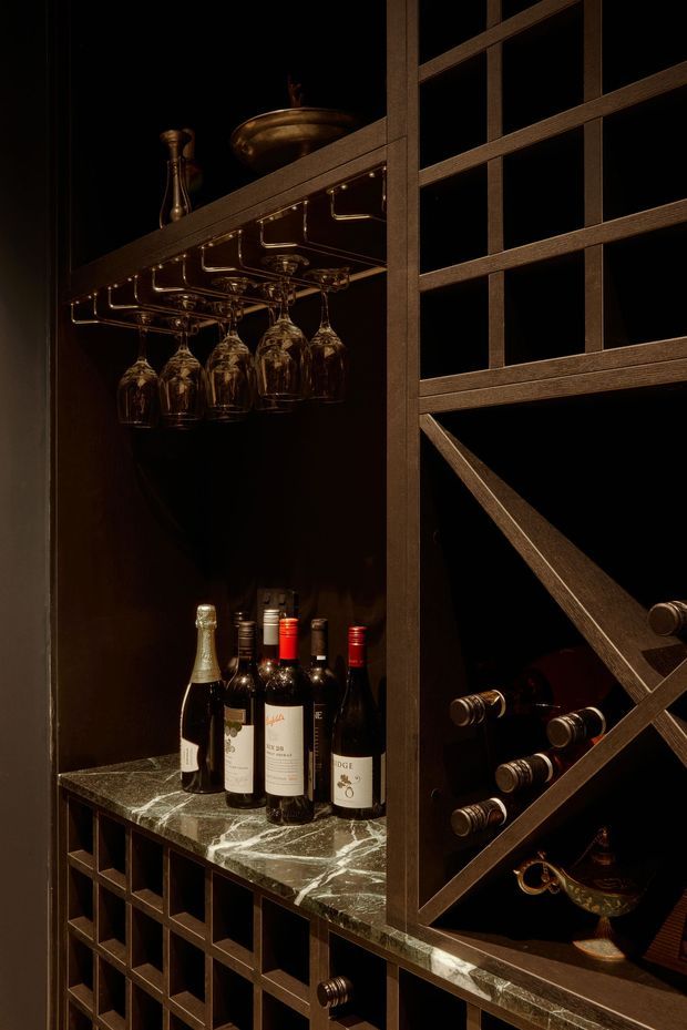 A dramatic, dark-toned built-in wine storage and bar display. The cabinetry features a bronze-brown metal grid with bottle racks, hanging stemware holders, dark shelving, wine bottles, and brass-toned decorative vessels. A polished veined stone countertop runs through the lower section, creating a refined cellar or home bar atmosphere.