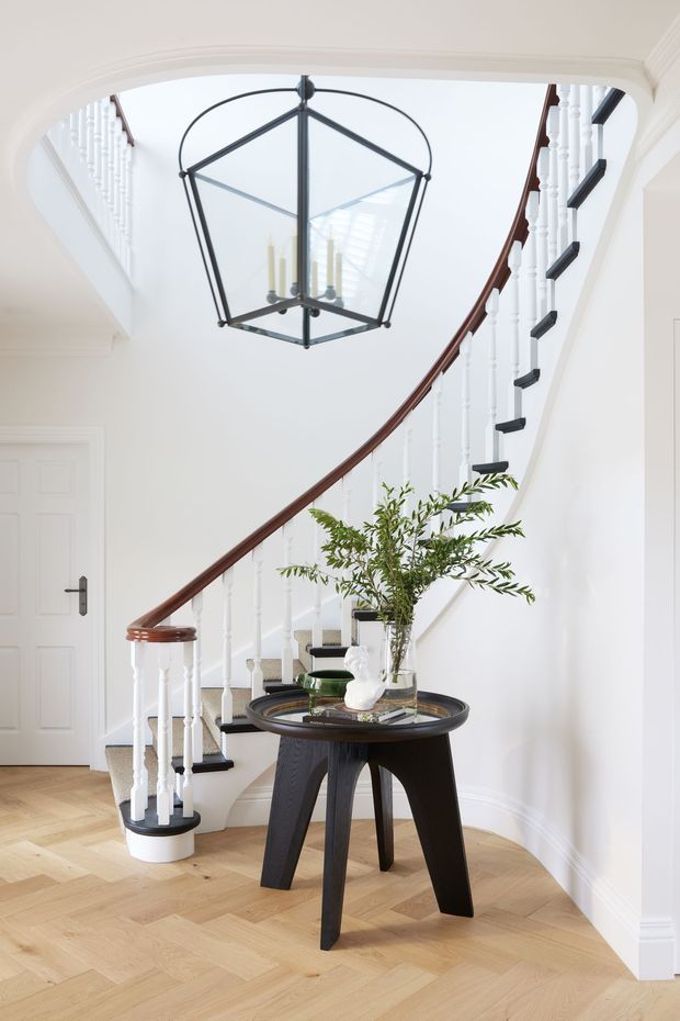Bright, refined entry hall featuring a sweeping curved staircase with white turned balusters, dark timber handrail, and black stair treads. A large black lantern pendant hangs beneath the stairwell opening, while a round dark timber console table with decorative objects and greenery sits on a pale herringbone timber floor. White walls, arched detailing, and traditional trims complete the elegant interior.