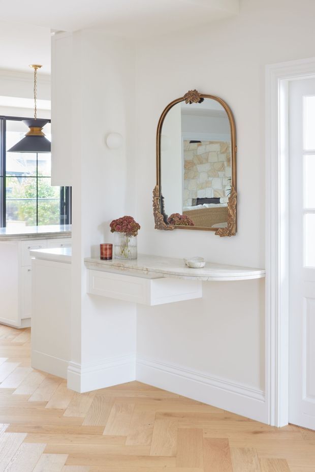 Bright, refined transitional entry hall with a floating white console and pale stone benchtop beneath an ornate antique-style arched mirror. The space features white walls and trim, light timber herringbone flooring, a decorative pendant visible through the opening to the kitchen, and abundant natural light. A white kitchen is partially visible to the left.