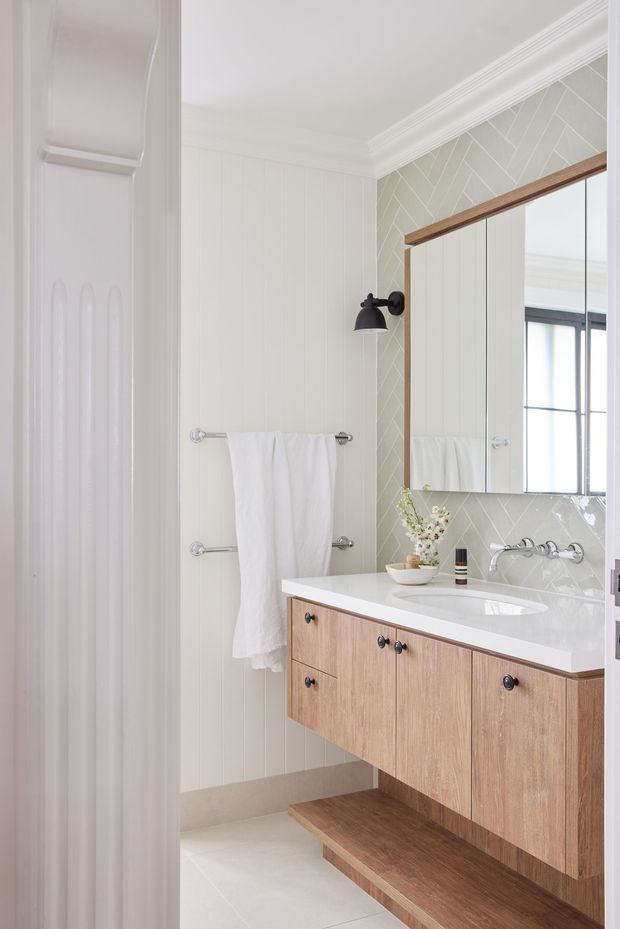 Bright, refined bathroom with white vertical wall panelling, pale patterned tile splashback, a large framed mirrored cabinet, white benchtop with integrated basin, and a floating light timber vanity with open lower shelf. Black wall sconce and chrome tapware provide understated contrast.