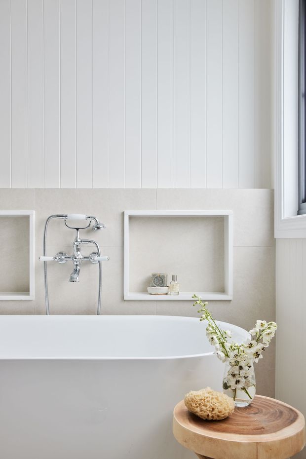 Bright, refined bathroom detail featuring a freestanding white bathtub with polished chrome wall-mounted fixtures, pale stone-look wall tiling, white vertical wall panelling, recessed display niches, and a white-framed window. A small round timber side table with flowers and bath accessories sits beside the tub.