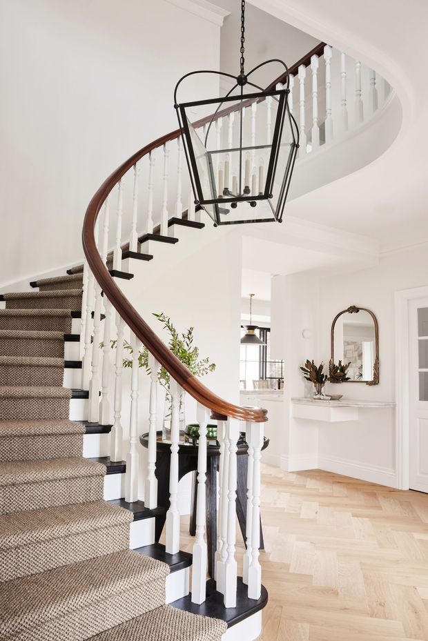 Bright, refined entry hall centred on a sweeping curved staircase. The staircase features white turned balusters, dark painted stair edges and newel details, a polished dark timber handrail, and a textured neutral carpet runner. A large black metal lantern pendant hangs above the stairwell. Pale herringbone timber flooring, white painted walls and trim, an arched brass-framed mirror, console shelf, and glimpses of the kitchen complete the elegant interior.