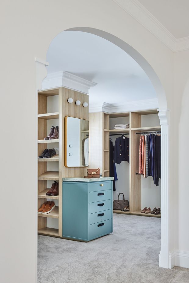 Bright, refined walk-in wardrobe viewed through a large white decorative archway. The room features floor-to-ceiling light timber shelving and hanging storage, a central blue-green drawer island with a stone benchtop, integrated mirror, brass-toned hardware, clothing rails, shoes, and soft grey carpet. White walls, ornate cornices, and classical architrave details create a polished traditional shell, while the cabinetry and island add a contemporary tailored feel.