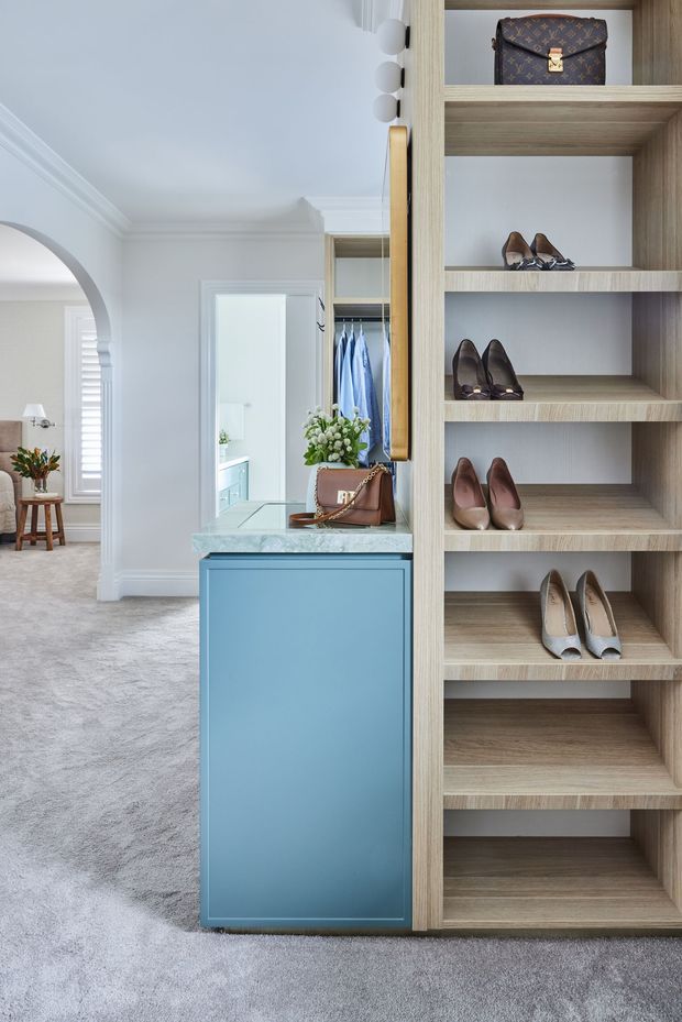 A bright, refined walk-in wardrobe or dressing area with floor-to-ceiling open shoe shelving in light wood, a blue built-in cabinet with a pale stone benchtop, and a mirrored panel. Clothes are visible in the wardrobe behind, while an adjoining elegant bedroom or sitting area can be seen through an arched opening. The room features white painted walls with decorative moulding, grey carpet, and abundant natural light.