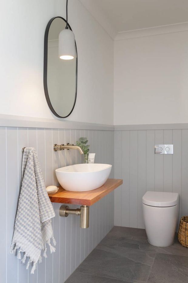 Bright, compact bathroom featuring grey vertical wall panelling, dark grey floor tiles, a wall-mounted timber vanity with a white vessel basin, brushed brass tapware, an oval black-framed mirror, pendant light, and a concealed-cistern toilet.