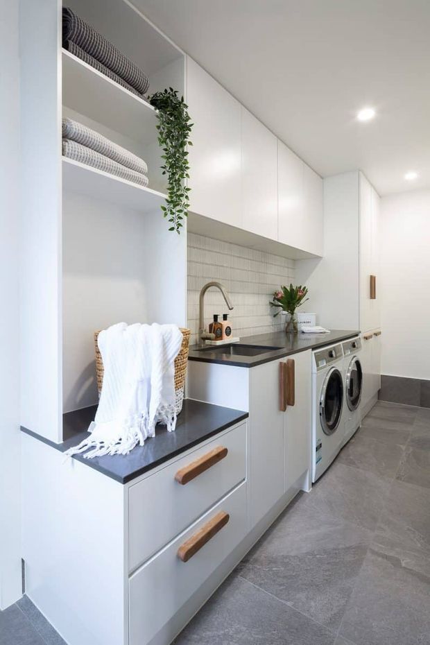 Bright, contemporary laundry room with extensive white built-in cabinetry, open shelving, integrated washer and dryer, a dark stone-look benchtop, and a textured light tile splashback. Warm timber drawer pulls, woven baskets, folded towels, and a trailing plant add natural accents. Large grey floor tiles and recessed ceiling lights complete the clean, functional space.
