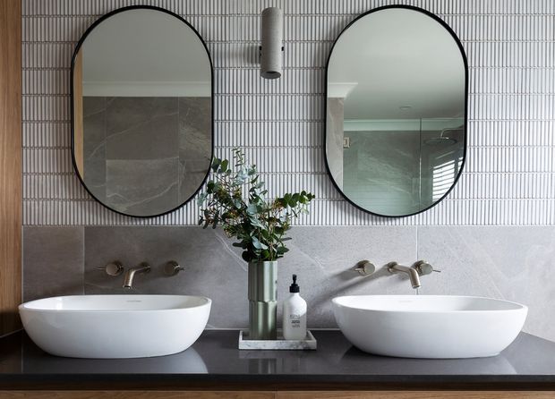 A refined double-vanity bathroom with two white vessel basins on a dark stone countertop, wall-mounted brushed metal tapware, large rounded black-framed mirrors, and a vertically ribbed light tile feature wall. Grey stone wall tiles and timber cabinetry add texture and warmth, with a small plant and bathroom accessories between the basins.