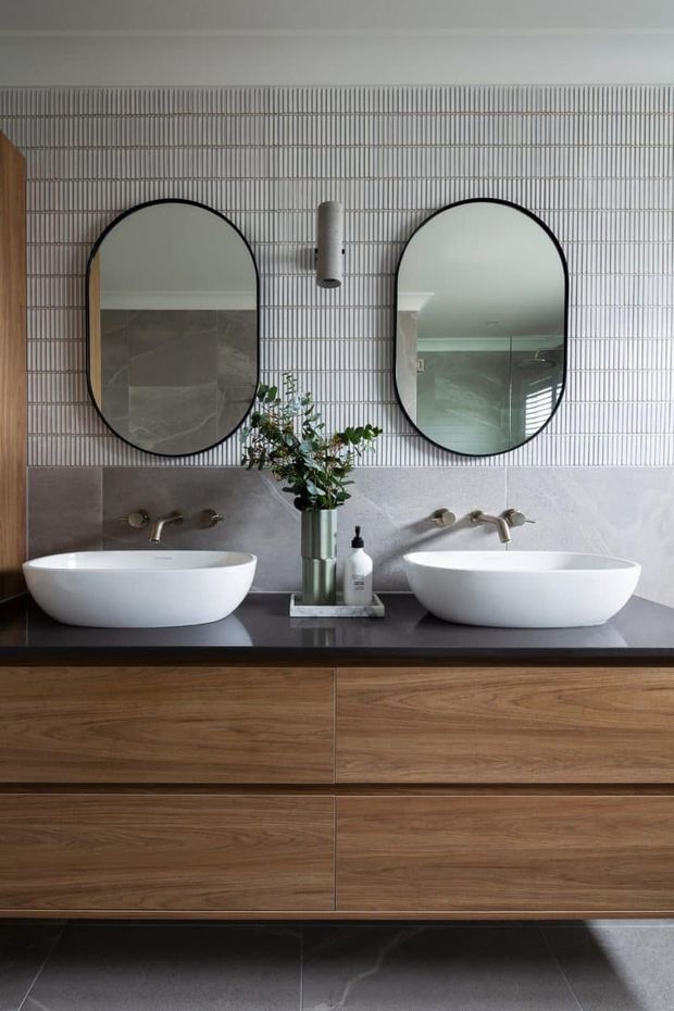 A refined double-vanity bathroom featuring two white vessel basins on a dark stone benchtop, a long timber vanity with integrated drawers, oval black-framed mirrors, wall-mounted brushed metal tapware, and a textured vertical wall tile backdrop. A small floral arrangement and toiletries sit between the basins.