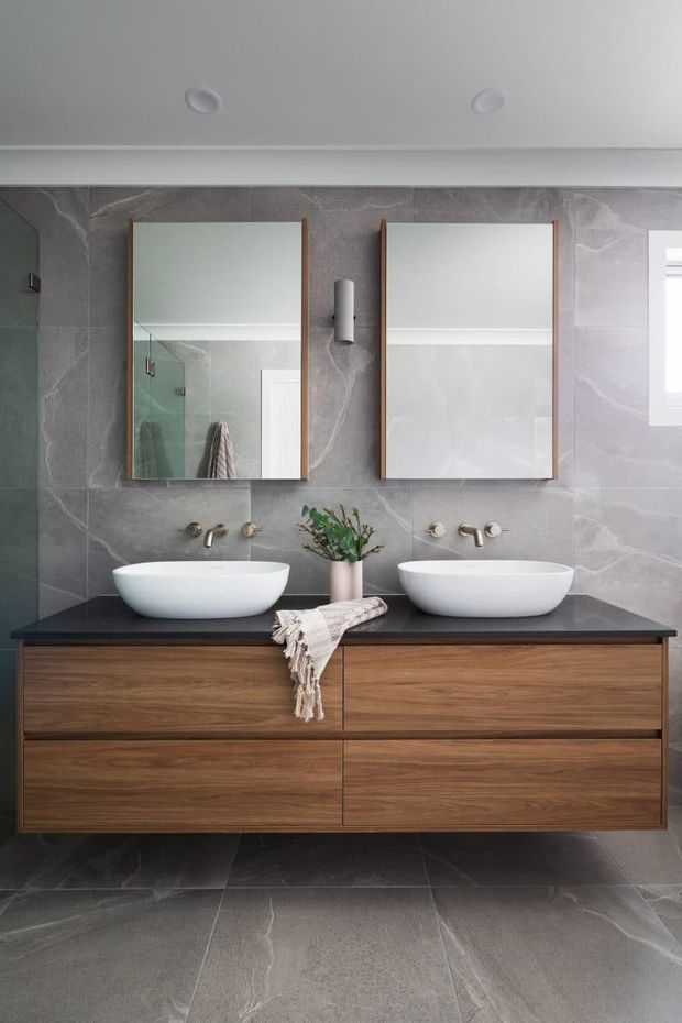 A contemporary bathroom featuring a wide floating double vanity with warm walnut-toned timber cabinetry, a dark stone benchtop, two white vessel basins, wall-mounted brushed-metal tapware, and twin rectangular mirrors. Large grey veined stone-look tiles cover the walls and floor, creating a refined, neutral-toned interior.