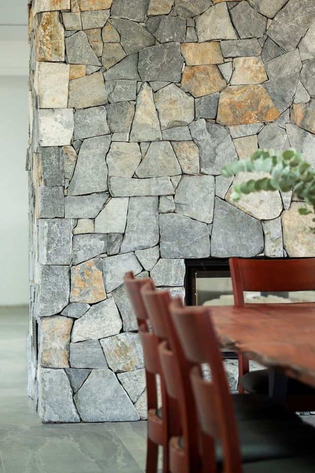 Interior detail showing an irregular natural stone feature wall surrounding a dark fireplace opening, with a timber dining table and chairs in the foreground. The stonework is predominantly grey with warm tan and rust-coloured variations, and the floor appears to be a smooth grey stone or concrete finish.