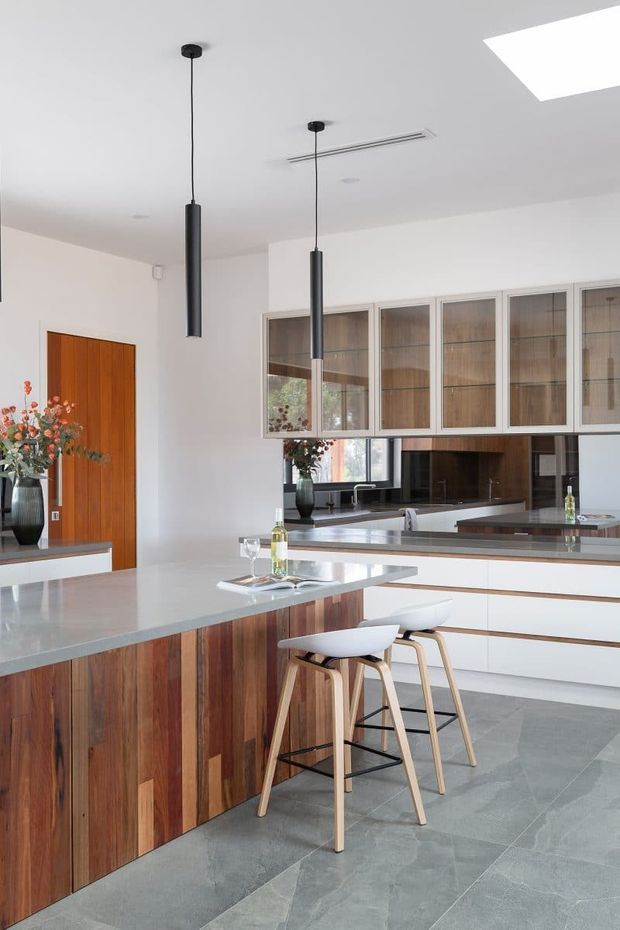 Bright contemporary kitchen featuring a large island with vertically grained timber-clad cabinetry, a pale stone benchtop, integrated white cabinetry, glass-fronted overhead cupboards, dark reflective splashback, and two slim black pendant lights. Two light timber bar stools sit at the island, with grey stone-look flooring and a timber door adding warmth.