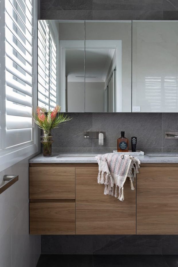 A refined contemporary bathroom vanity with a wall-mounted timber-grain cabinet, pale stone benchtop, dark large-format tiled splashback and matching dark floor tiles. A wide mirrored cabinet spans the wall above the vanity, while plantation shutters provide filtered daylight at the side. The space includes a modern basin and tapware, understated toiletries, and a textured neutral towel draped over the counter.