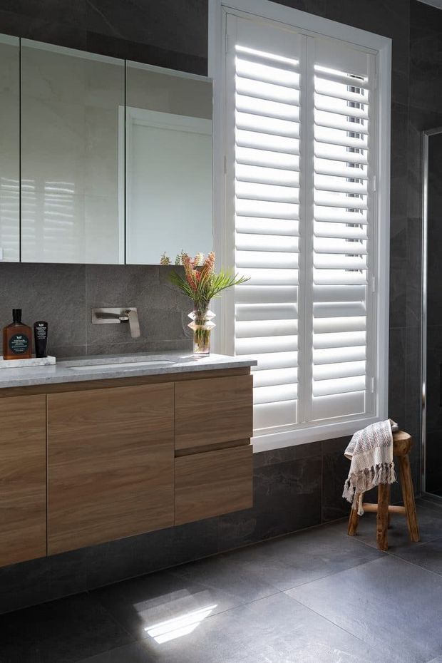 Contemporary bathroom featuring a floating walnut-toned vanity with integrated drawers, a pale stone benchtop, wall-mounted tapware, mirrored cabinetry, dark grey tiled walls and floor, and a large white plantation-shuttered window. A small wooden stool with a draped towel sits beside the window.