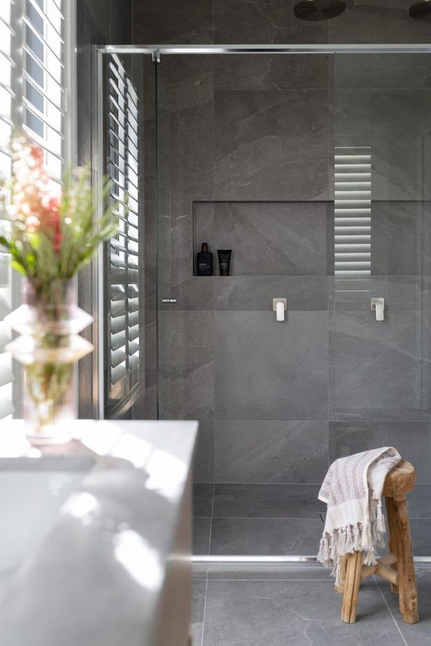 A refined contemporary bathroom centered on a large frameless glass shower enclosure. The shower walls and floor are clad in large-format dark grey stone-look tiles, with a recessed niche, wall-mounted controls, and a narrow vertical louvred window providing filtered daylight. A white freestanding or built-in bathtub occupies the foreground beside a bright window with plantation shutters. A simple timber stool with a draped textured towel adds warmth.