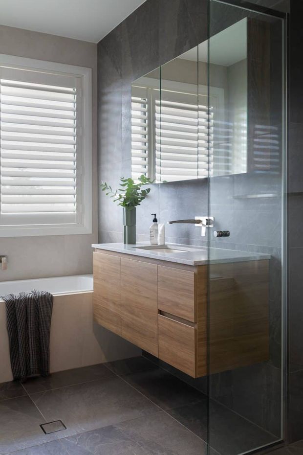 Contemporary bathroom with a floating timber vanity, white stone benchtop, large mirrored wall cabinets, grey tiled walls and floor, and a frameless glass shower enclosure. A bathtub sits beneath a shuttered window, with restrained greenery adding a natural accent.