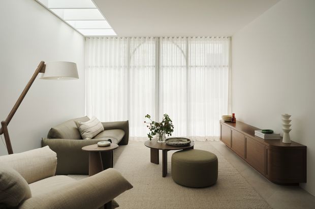 A bright, refined living room with floor-to-ceiling sheer curtains, a large skylight or clerestory-like ceiling panel, neutral upholstered seating, nested round coffee tables, an olive-toned ottoman, and a low dark timber media console. The space has clean contemporary lines and a calm, minimalist aesthetic.