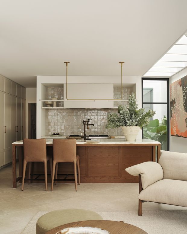 Warm, refined open-plan kitchen and living area with a central timber island, integrated cabinetry, pale stone-look flooring, textured tiled splashback, brass pendant lighting, upholstered bar stools, and a sculptural cream lounge chair. A large skylight and glazed opening bring in abundant natural light, while artwork and greenery add soft visual accents.