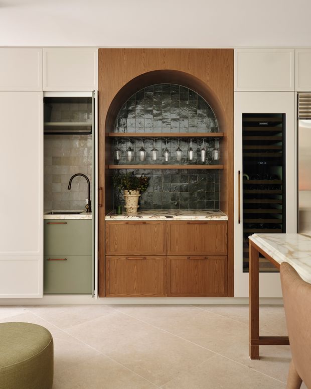 A refined kitchen interior focused on a built-in drinks and glassware display. The central joinery is crafted in warm timber with an arched recess, dark textured tile backsplash, open timber shelves, drawers, and a stone benchtop. Cream cabinetry surrounds the feature unit, with a compact sink zone to the left and a tall integrated wine fridge to the right. A marble-look island with timber stools is partially visible in the foreground. The space has a warm, sophisticated, contemporary-traditional feel.