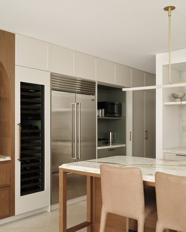 A refined contemporary kitchen with a large marble island and timber base, upholstered dining stools, full-height cream cabinetry, stainless-steel refrigerator, integrated wine fridge, and a dark built-in beverage or prep area. A slim brass linear pendant hangs above the island, while open shelving and a tiled wall are visible to the right. The palette is warm, neutral, and softly luxurious.