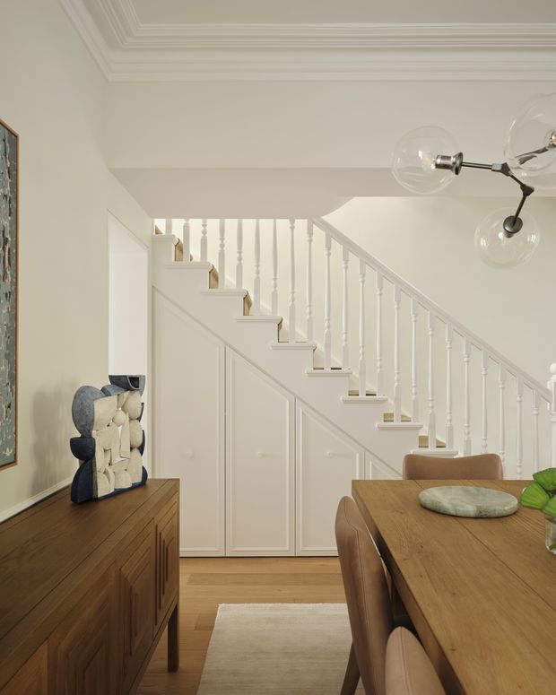 A refined dining area and hallway beneath a traditional staircase. The room features warm timber flooring and dining furniture, a timber sideboard, pale painted walls, detailed decorative ceiling cornices, and a white staircase with turned balusters and paneled under-stair storage. A sculptural pendant light hangs above the dining table, creating a warm, elegant interior.
