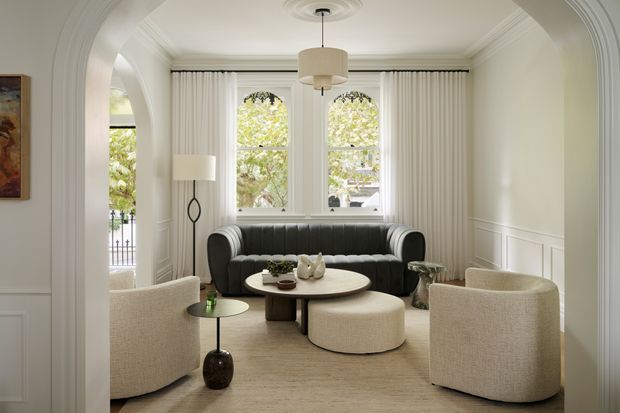 Elegant, light-filled living room with a refined traditional architectural shell. The room features a large arched opening with layered decorative trim, white wall panelling and moulded cornices, tall arched windows dressed with full-height curtains, and a central pendant light. A dark curved sofa is paired with two rounded cream boucle armchairs, a round coffee table, an ottoman, and a small side table on a neutral rug. The overall composition is calm, polished, and softly contemporary with classic detailing.