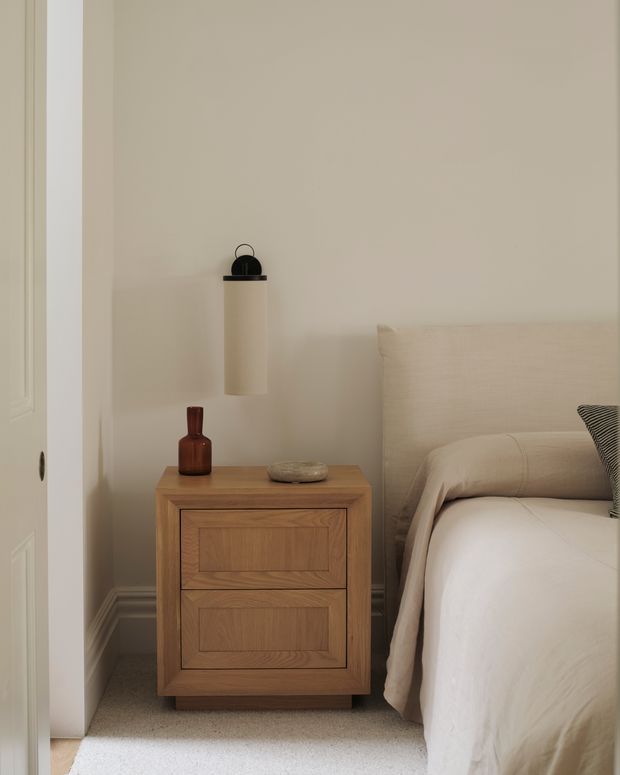 A softly lit bedroom vignette showing a warm natural timber bedside table with recessed-panel drawers, a neutral upholstered bed with cream bedding, beige walls, carpeted flooring, and understated decorative objects. A wall-mounted cylindrical pendant light and partially visible door frame the scene. The overall composition is calm, warm, and minimalist.