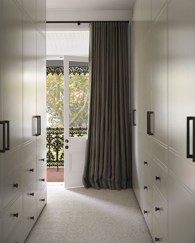 A narrow, symmetrical walk-in wardrobe lined with white built-in drawer cabinetry on both sides, featuring dark rectangular hardware. A tall pleated charcoal curtain frames an open white door leading to a small balcony or exterior landing with ornate black metal balustrades and leafy trees beyond. The room has pale carpet, white walls, and abundant natural light.
