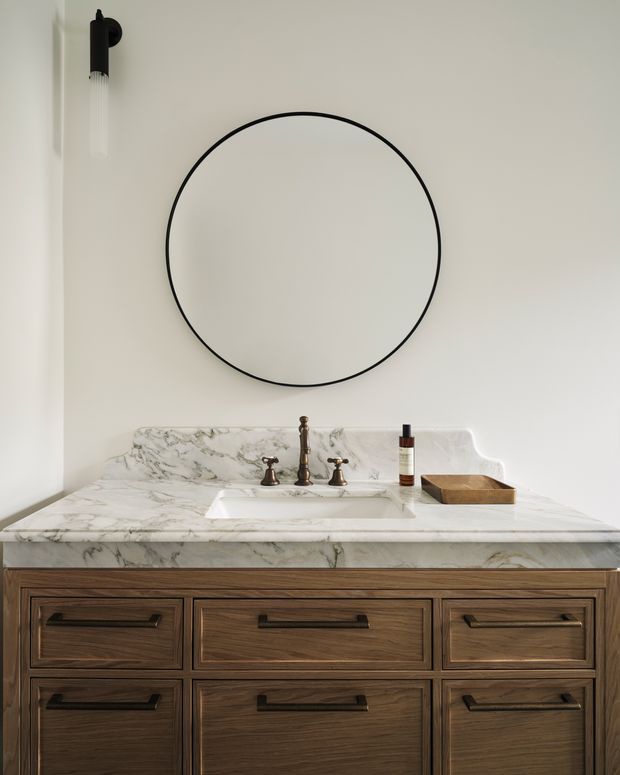 A refined bathroom vanity vignette featuring a dark timber vanity with black hardware, a white marble benchtop and integrated backsplash, a rectangular undermount basin with traditional chrome taps, and a large round black-framed mirror. A minimalist wall sconce and small timber tray complete the calm, classic composition.