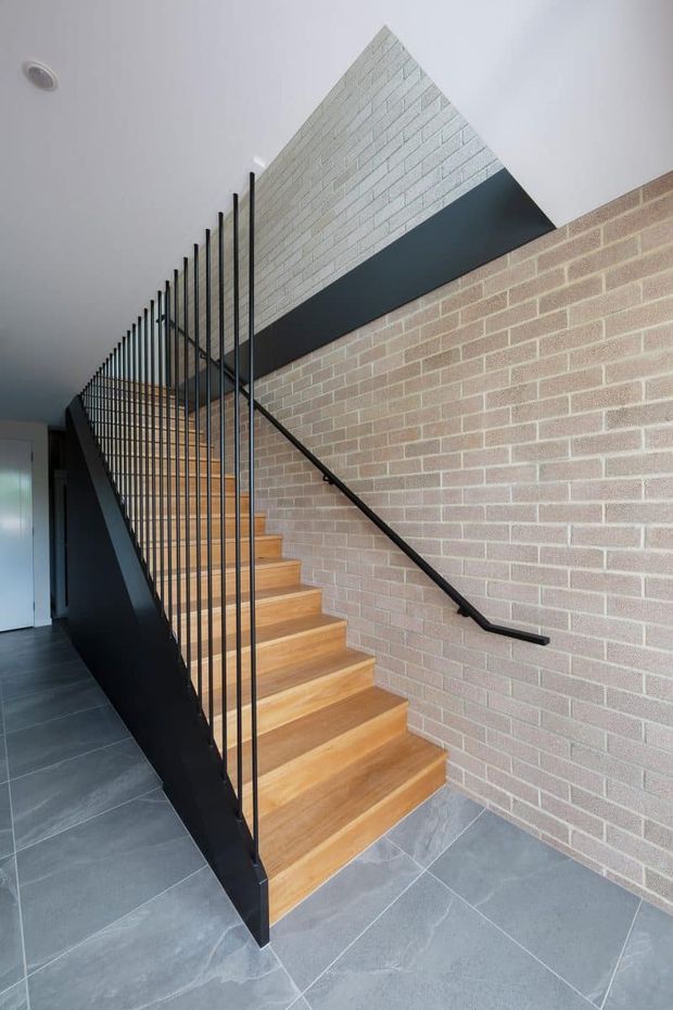 A contemporary interior stairway with warm timber treads, slim black metal balustrades, and a black handrail. The staircase is bordered by exposed light brick walls, with large grey floor tiles and a white ceiling creating a crisp, modern material palette.