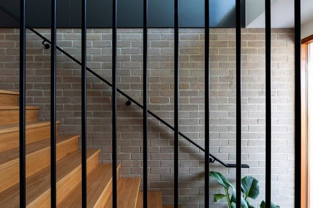 A contemporary stairway with warm timber treads ascending beside a grey exposed brick wall. Slim black vertical metal balusters form a strong screen along the staircase, complemented by a black handrail. A small indoor plant and a partial window add natural light and greenery.