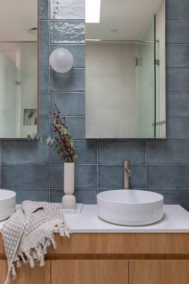 Contemporary bathroom vanity featuring a white vessel basin, brushed metal tapware, timber-look cabinetry, and large blue-grey rectangular wall tiles. A mirrored cabinet, glass shower screen, recessed shelf, and simple decorative vase complete the clean, modern composition.