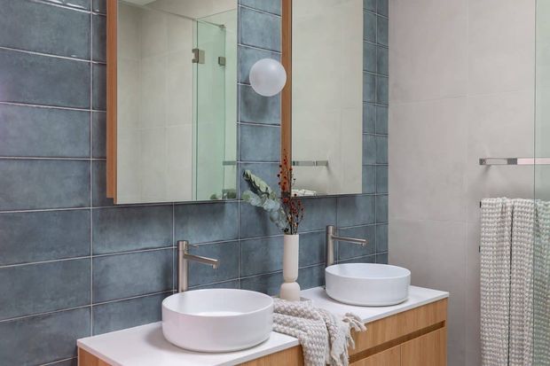Contemporary double-vanity bathroom with two white vessel basins, brushed metal wall-mounted taps, a timber vanity with a pale benchtop, large mirrors, and a frameless glass shower enclosure. The walls combine dark blue-grey rectangular tiles with pale smooth tiles, creating a refined, cool-toned scheme.