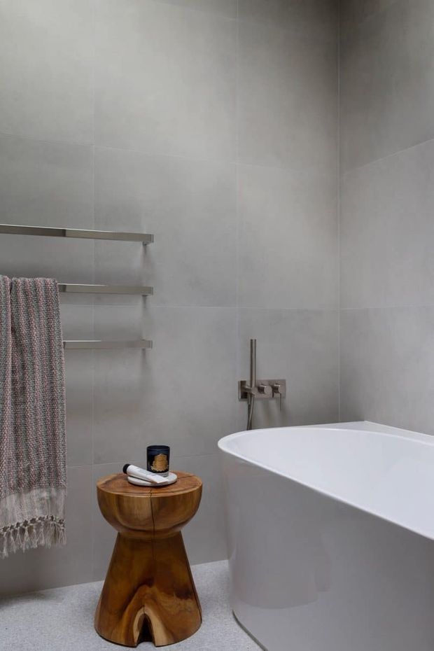 A refined minimalist bathroom featuring a freestanding white bathtub, large pale grey wall tiles, brushed metal wall-mounted tapware, slim towel rails, and a sculptural warm timber stool. A textured neutral towel adds softness to the otherwise calm, monochromatic space.