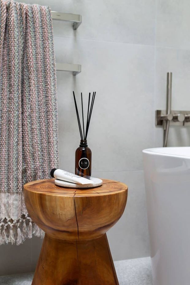 Close-up of a contemporary bathroom featuring a freestanding white bathtub, pale tiled walls, brushed metal bath fittings, open wall shelves, and a rustic solid timber pedestal stool or side table. A textured pink, grey, and cream shower curtain and reed diffuser add softness and colour.