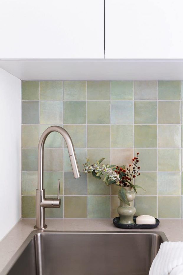 Close-up view of a refined kitchen sink area featuring a brushed metal gooseneck faucet, deep stainless-steel undermount sink, pale stone benchtop, and a pastel green and cream tiled splashback. A small decorative vase and soap dish sit on the counter.