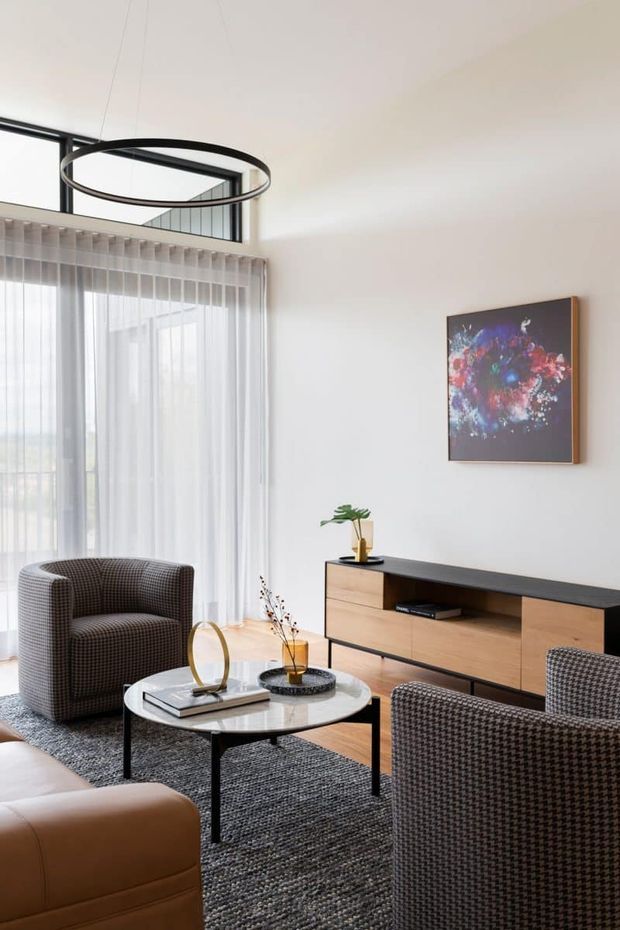 Bright contemporary living room with a high ceiling, full-height sheer curtains, black-framed clerestory glazing, and a slim circular pendant light. The room features a sculptural upholstered armchair, brown leather seating, patterned lounge chairs, a round marble-look coffee table, dark textured rug, and a low timber-and-black media console with artwork above.